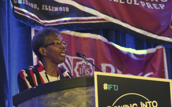 An older Black woman with glasses stands behind a podium with an "Ignatian Family Teach-In for Justice" poster on the front