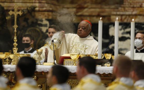 A Black man wearing a red zucchetto disperses incense behind a large altar