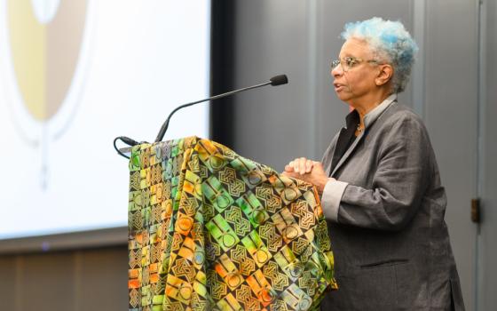 An older Black woman with blue streaksin her hair speaks at a podium covered in a brightly colored cloth