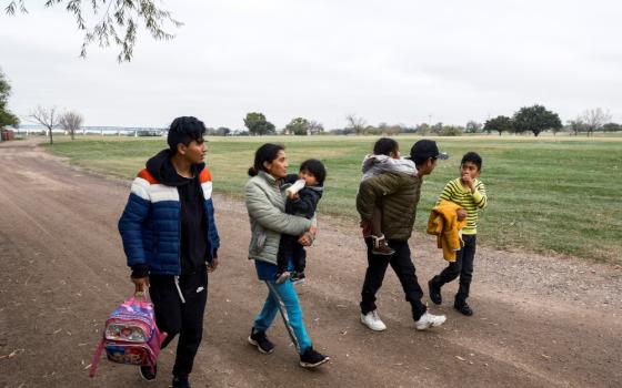 A family with adults carrying children walks along a dirt road next to a grass fiedl