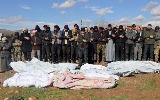 A group of people stand outside around bodies fully wrapped in cloth