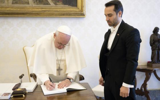 Pope Francis writes in a notebook on a desk. Next to him stands a man in a tuxedo