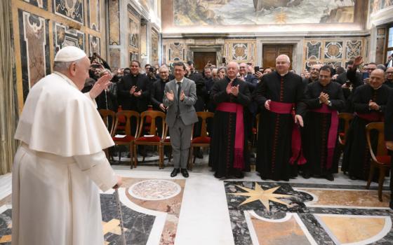 Pope Francis stands in front of and holds his hand in a blessing gesture towards a large audience of men, mostly clerics