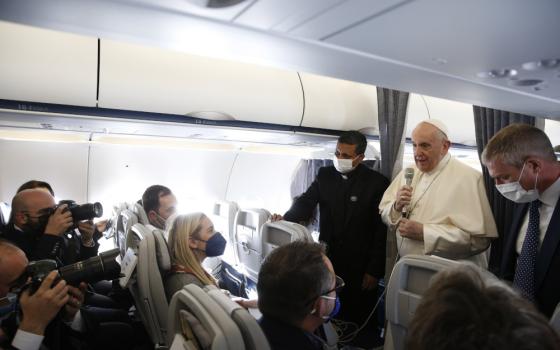 Pope Francis stands in the middle of a plane and speaks with masked journalists who surround him