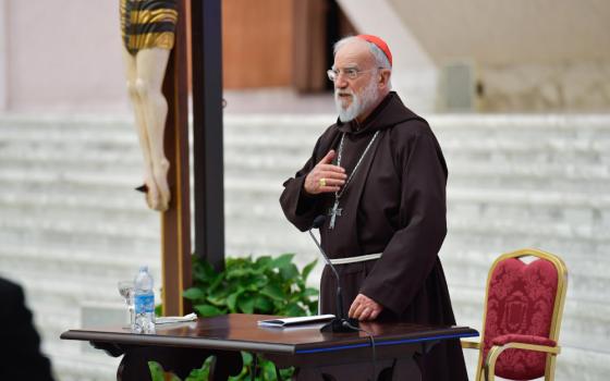 An older white man in a red zucchetto, pectoral cross and brown tunic with a chord speaks next to a crucifix