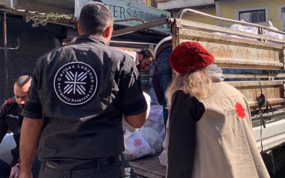 Two people whose backs face the camera wear Caritas jackets. They face a truck.