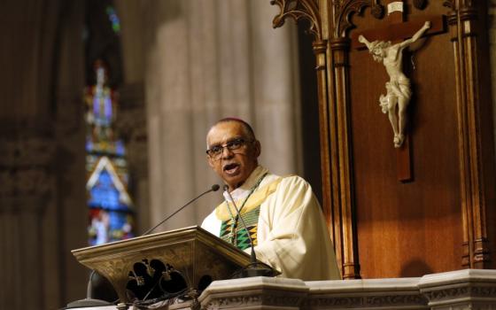 A light-skinned Black man speaks behind a lecturn and in front of a crucifix