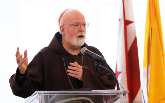 An older white man with a beard and glasses wears a red zucchetto and a brown tunic