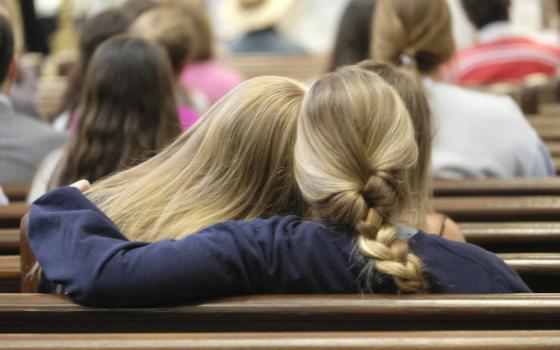 A person with long blond hair leans on a person with a blond braid, who wraps her arm around the other person. They sit in a church pew.