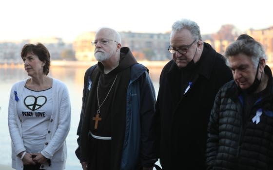 An older white man wears a brown robe and a wooden pectoral cross as he stands between two men and a woman