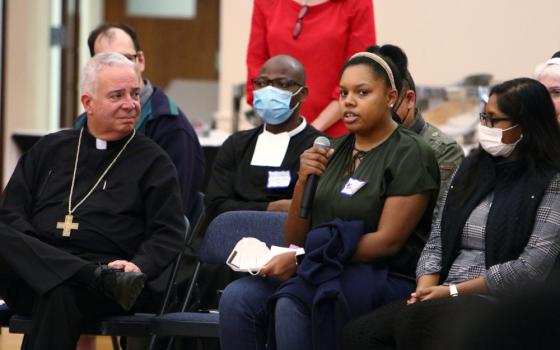 A man wearing a black cassock and a pectoral cross looks on as a young Black woman speaks into a microphone, while surrounded by other people in chairs, some masked