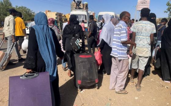 A group of people with suitcases gather outside white buses. Some women wear headscarves