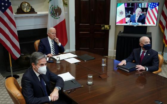 Two men in suits and masks sit on either side of Joe Biden, also in a suit, at a long table. A man with a Mexican and US flag behind him is on a video screen.