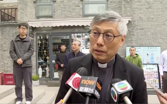 In this image from a video shot by Hong Kong broadcaster TVB, Hong Kong Bishop Stephen Chow talks to media during his visit to Beijing, with unidentified men standing behind him, on Thursday, April 20, 2023. Chow said in a historic trip to Beijing that he hopes the dioceses of the two cities will have more exchanges and cooperation, local media reported Thursday amid signs of Sino-Vatican strains. (TVB via AP)