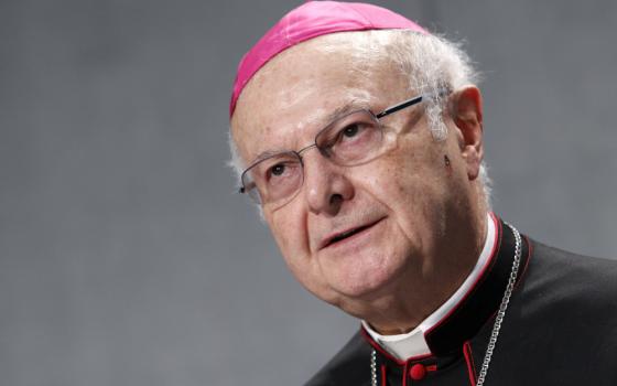 An older white man wearing glasses wears an amaranth zuchetto along with a black cassock