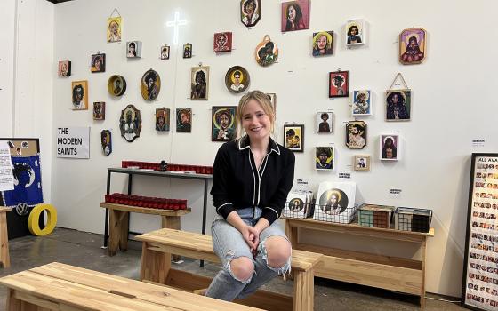 Gracie Morbitzer, a young white woman in ripped jeans and a black top, sits on a bench in her studio