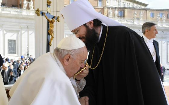 Pope Francis, sitting, leans forward to kiss something hanging by a chain around a man's neck, who wears a black robe and a white koukoulion