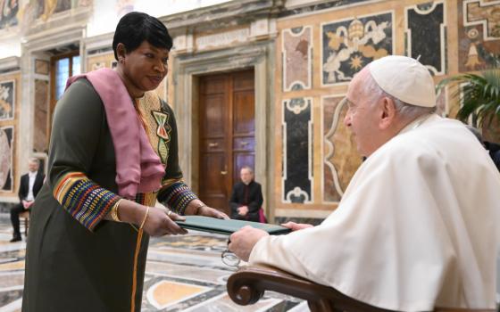 A Black woman who wears a olive green and multi-colored dress hands a folder to a seated Pope Francis