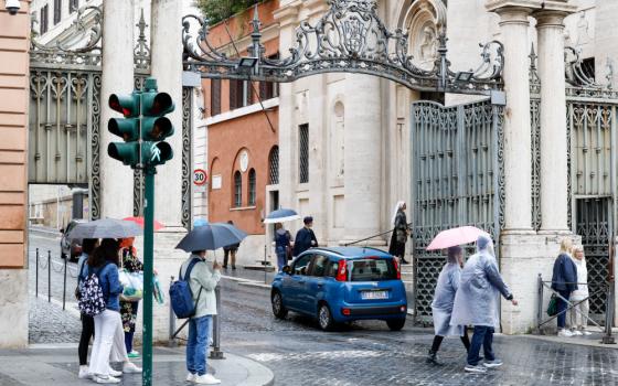 A small clue car drives through an ornate wrought iron gate. People walk by with umbrellas and rain ponchos.