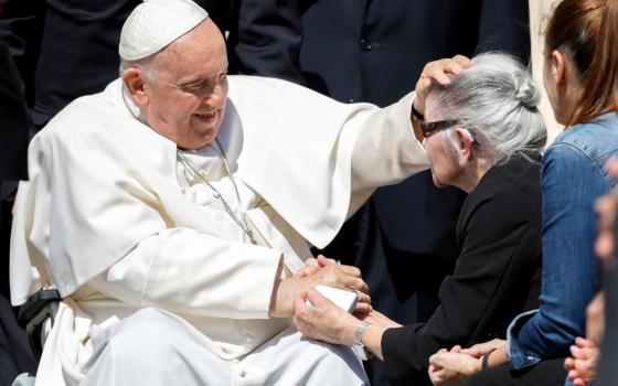 Pope Francis, sitting in his wheelchair, places a hand on the head and hands of an older white woman with a hearing aid