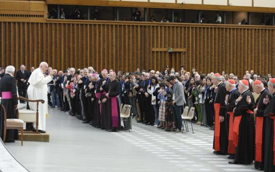 Pope Francis stands and raises his hand in front of a crowd that includes people dressed in the garb of cardinals, bishops and laypeople