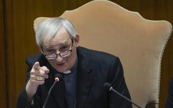 A man in a clerical collar and jacket speaks into a microphone and points his finger