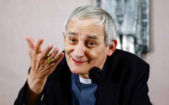 An older white man wearing a blue shirt, black jacket and clerical collar speaks into a microphone