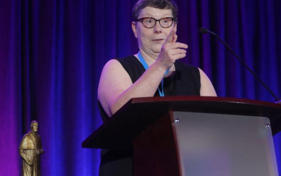 A white woman with short hair and glasses stands behind a podium and points as she is speaking