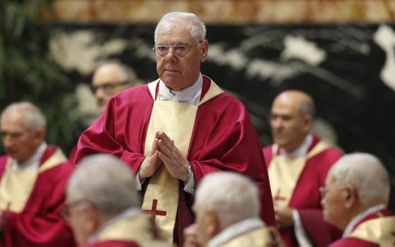 An older white man wears a red chasuble with a gold piece down the middle with a red cross on it. He stands with his hands together in front of him.