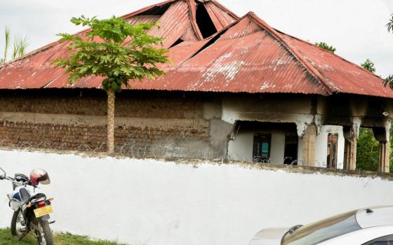 A building with an orange roof and brown brick sides shows black marks and gaps in the ceiling. It stands behind a white wall with barbed wire.