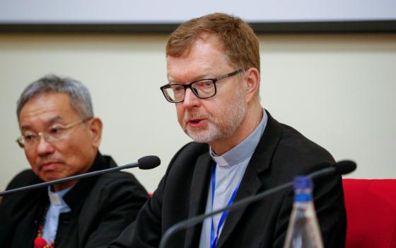 A white man wearing dark rimmed glasses and a clerical collar with a blue shirt and black suit speaks into a microphone