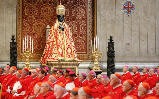 A black statue of St. Peter wears a red and gold robe and a red and gold dome-like tiara while towering over a large group of cardinals and bishops