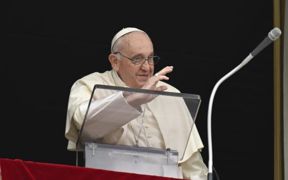 Pope Francis raises his hand to wave while standing behind a clear lectern and a microphone