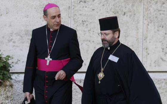 A man wearing a violet zucchetto stands next to another man wearing a skufia hat