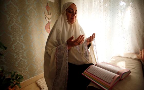 A woman wearing a waist-length hijab lifts her hands, palms raised, while sitting in front of a book with Arabic writing