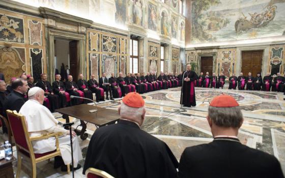 A man wearing a violet zucchetto stands in the middle of a room covered in frescos as he addresses Pope Francis and other bishops and cardinals