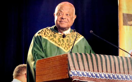 A Black bald man wears green and gold vestments and speaks from behind a podium