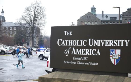 A sign that says "The Catholic University of America founded 1887 In Service to Church and Nation" is visible in front of a parking lot with police cars.