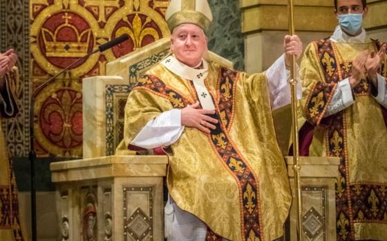 A white man wears a gold mitre and vestments and sits in an ornate chair holding a crozier