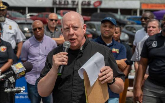 A white man wearing a clerical collared shirt speaks into a microphone as men stand behind him