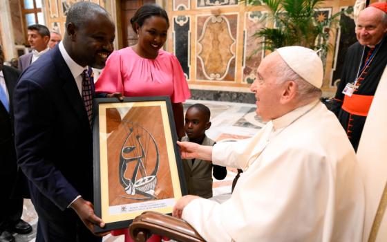 A Black man wearing a suit hands an an African painting of a musician to Pope Francis