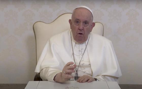 Pope Francis sits in a white backed chair against a white patterned wallpaper wall and speaks