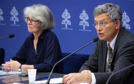 An older white man in a suit speaks into a microphone while seated at a table where an older white woman is present