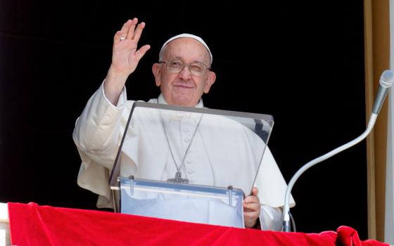 Pope Francis raises his hand to wave as he stands behind a clear lectern. There is a red tapestry over his window ledge.