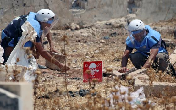 Two people wearing white helmets and light blue jackets crouch on the ground next to a red sign with a skull and crossbones