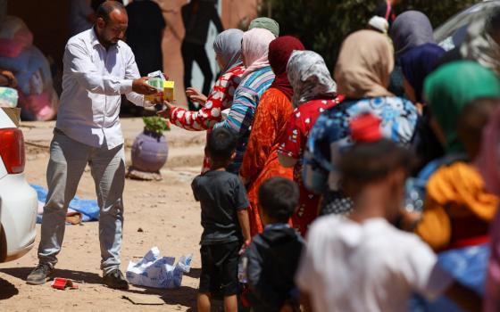 A man hands small boxes to a line of women dressed in colorful dresses and head scarves