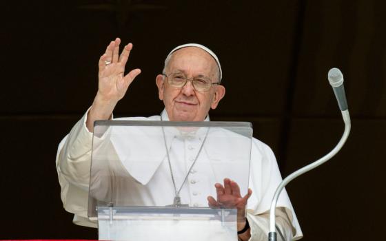 Pope Francis raises his hand to wave with the other hand on his clear lectern. A microphone is next to him.