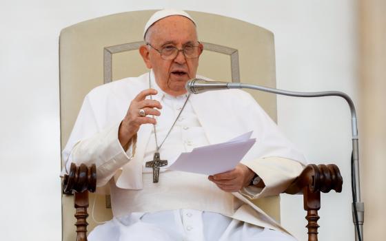 Pope Francis speaks into a microphone while sitting in a large cream chair and holding a piece of paper