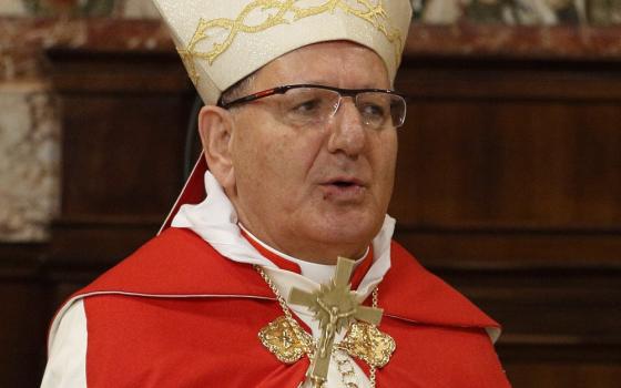 A light-skinned man wears a mitre, glasses, and red and white vestments and carries a gold crucifix