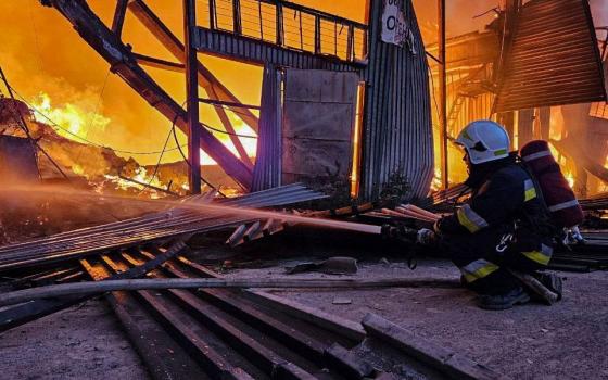 A person in a white helmet sprays water into a landscape full of fire and a burned out building